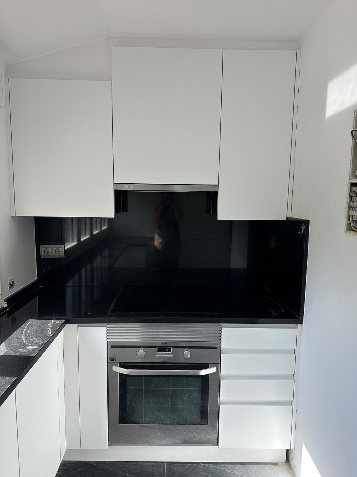 Cocina moderna y compacta con armarios blancos, salpicadero negro, horno de acero inoxidable y encimera en una habitación pequeña y luminosa
