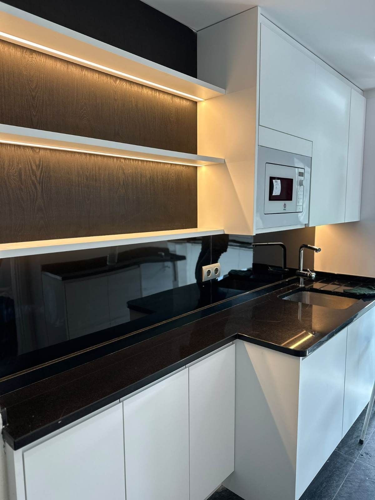 Cocina moderna con encimera negra, armarios blancos e iluminación cálida bajo los estantes