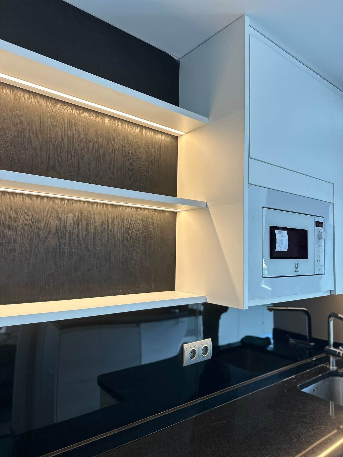 Cocina moderna con estantes abiertos iluminados, encimera negra y horno empotrado en la pared