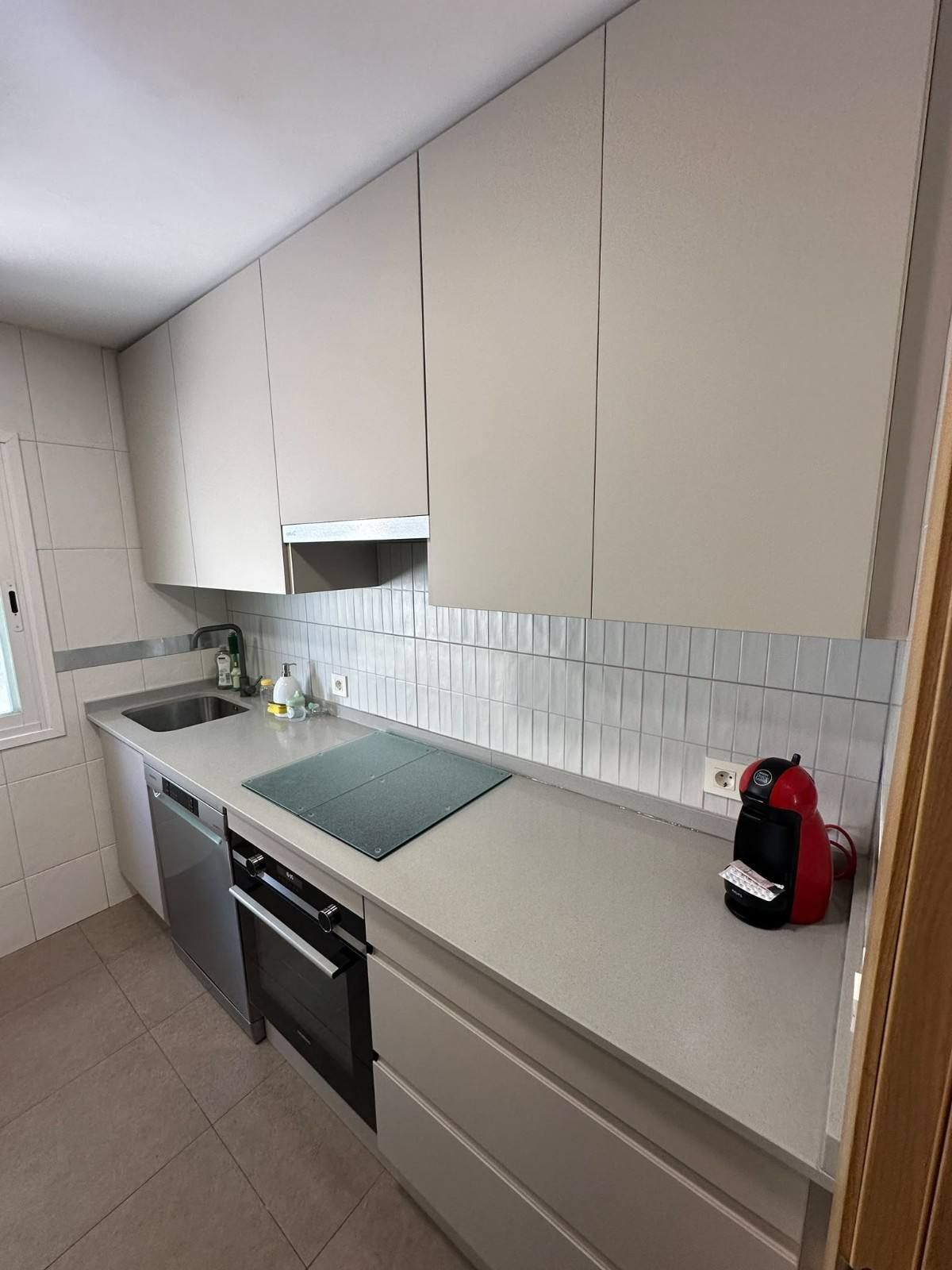 Cocina moderna con armarios beige, encimeras blancas, horno empotrado, fregadero y cafetera negra