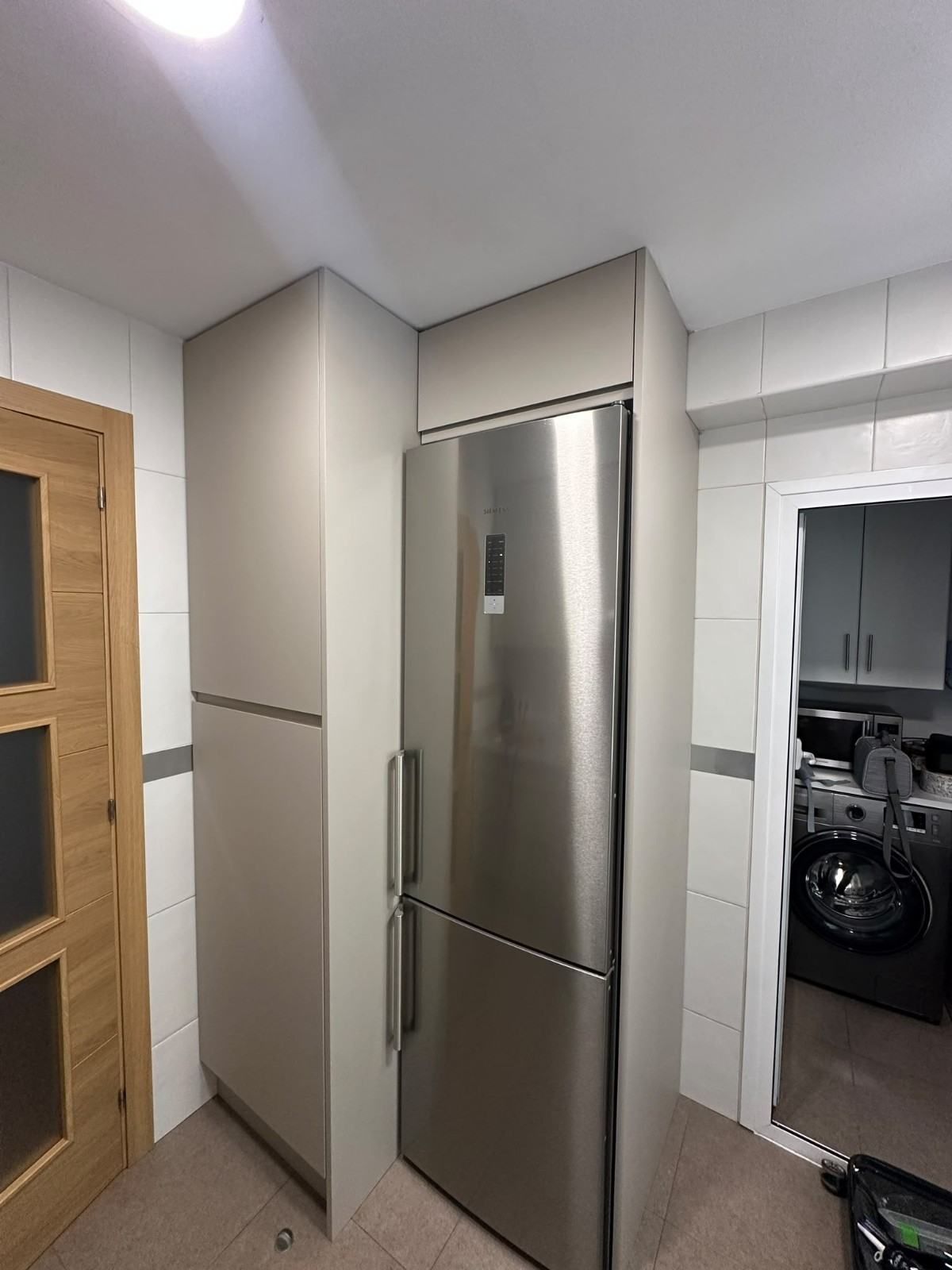 Refrigerador de acero inoxidable en un pasillo estrecho de cocina, con paredes de azulejos blancos y un panel de puerta de madera.