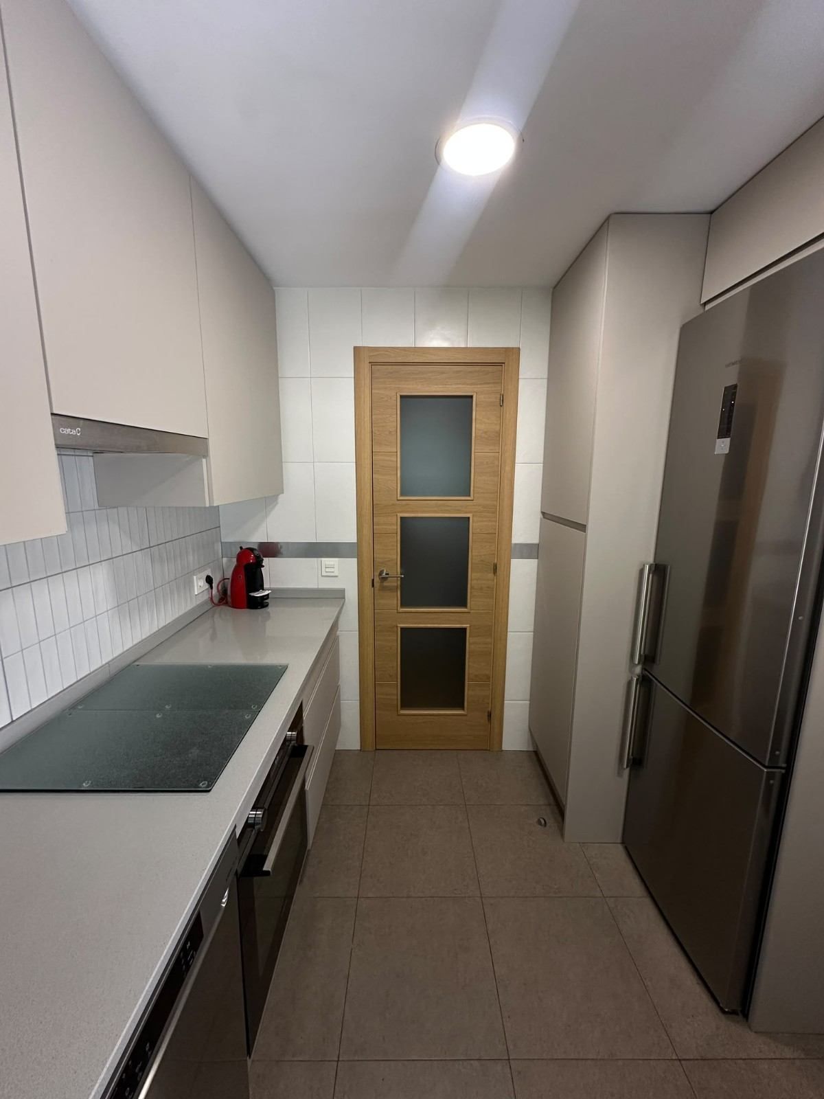 Cocina moderna y estrecha, con armarios blancos, frigorífico de acero inoxidable y una puerta de madera al fondo.