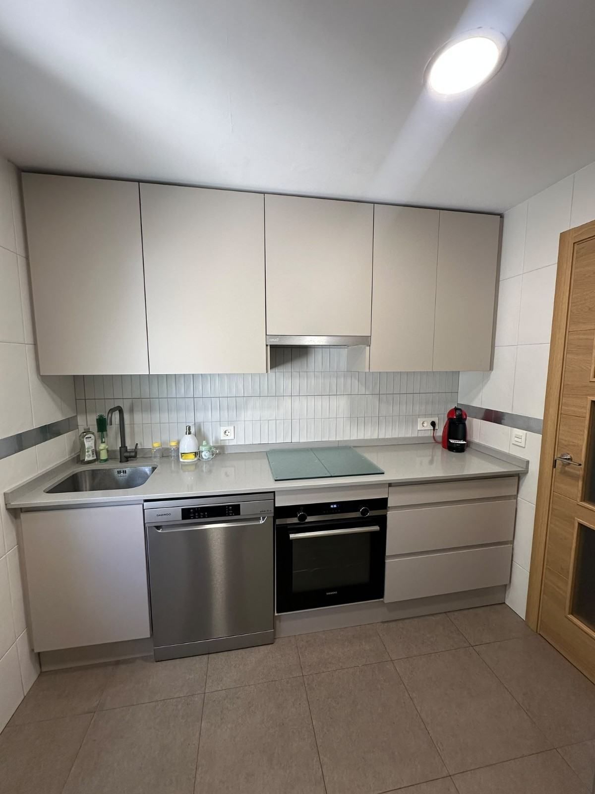Cocina moderna y compacta, con armarios beige, electrodomésticos de acero inoxidable, suelo de baldosas y una puerta de madera.