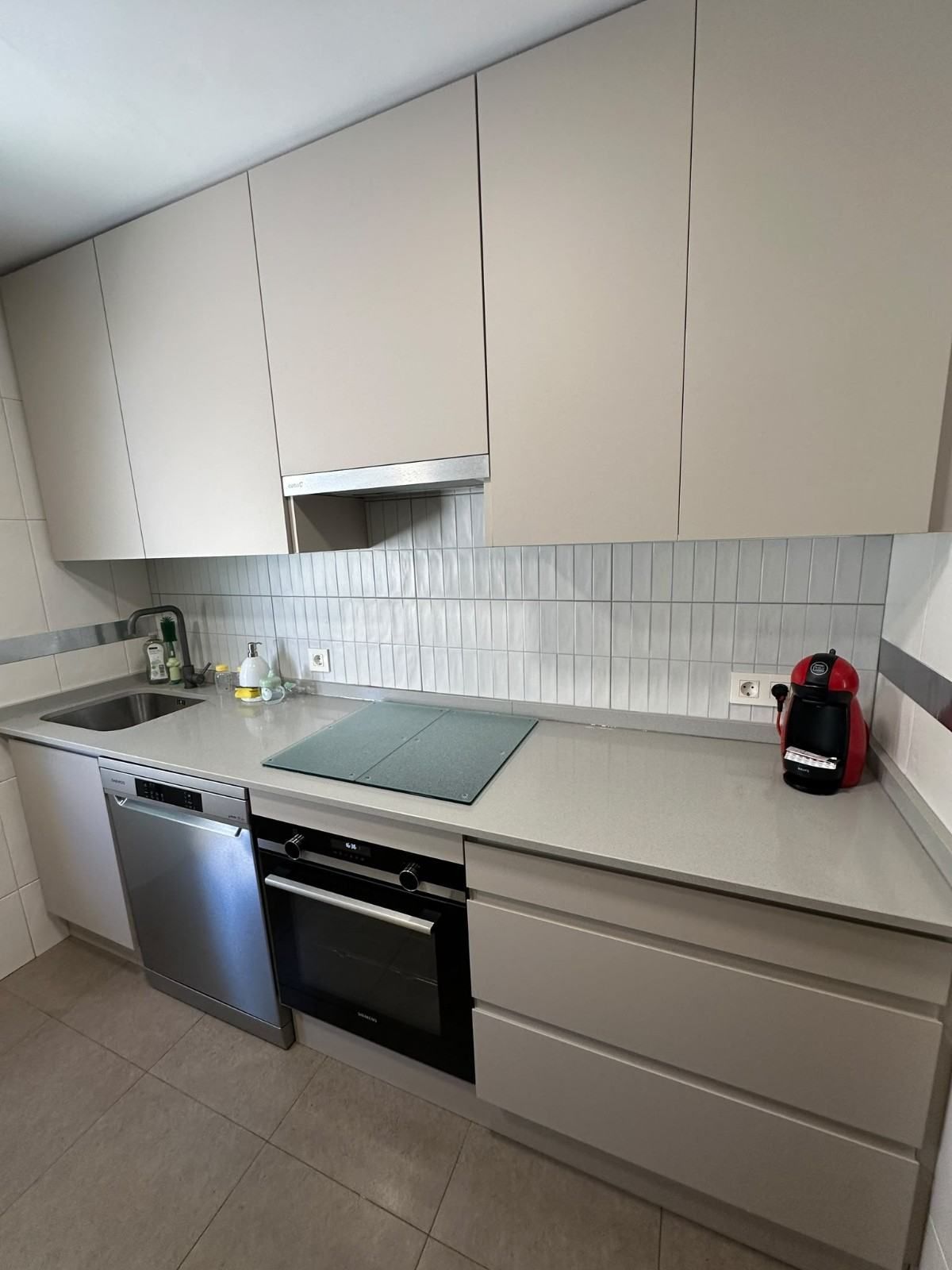 Cocina moderna con muebles beige, salpicadero blanco, lavavajillas de acero inoxidable, horno y electrodomésticos de encimera
