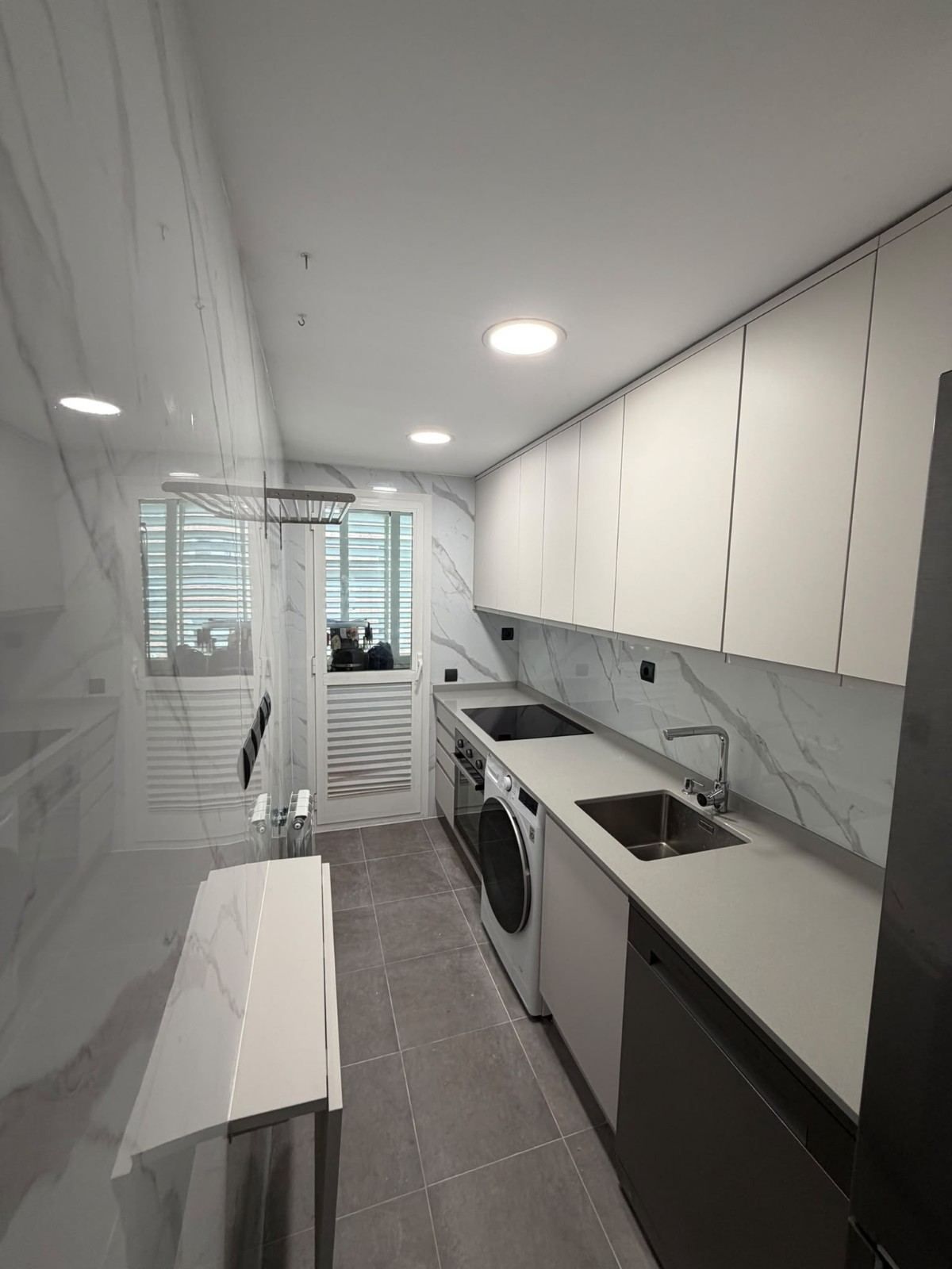 Cocina estrecha tipo pasillo, blanca, con lavadora, fregadero, armarios, suelo de baldosas grises y una pared del fondo con ventanas