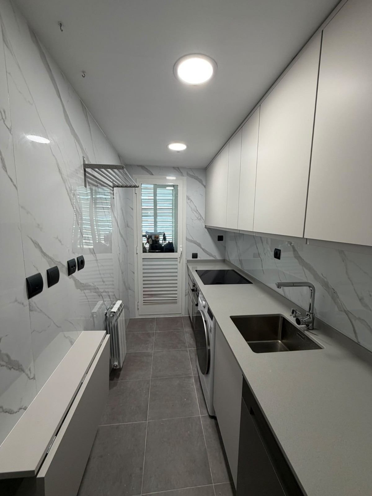 Cocina estrecha y blanca en forma de pasillo, con paredes de mármol, suelo gris, fregadero, armarios y una puerta con ventana al fondo.