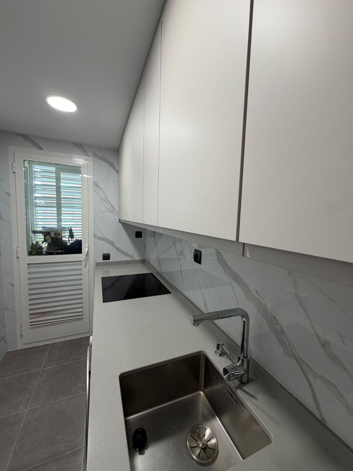 Cocina moderna y estrecha con armarios blancos, salpicadero de mármol, fregadero doble y una ventana al fondo.