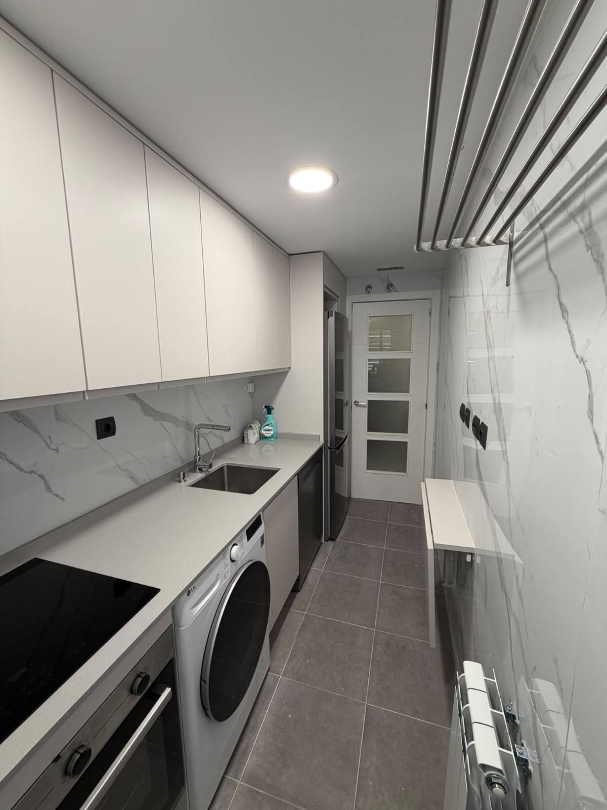 Cuarto de lavado estrecho y blanco con lavadora, secadora, encimera, armarios y suelo de baldosas.