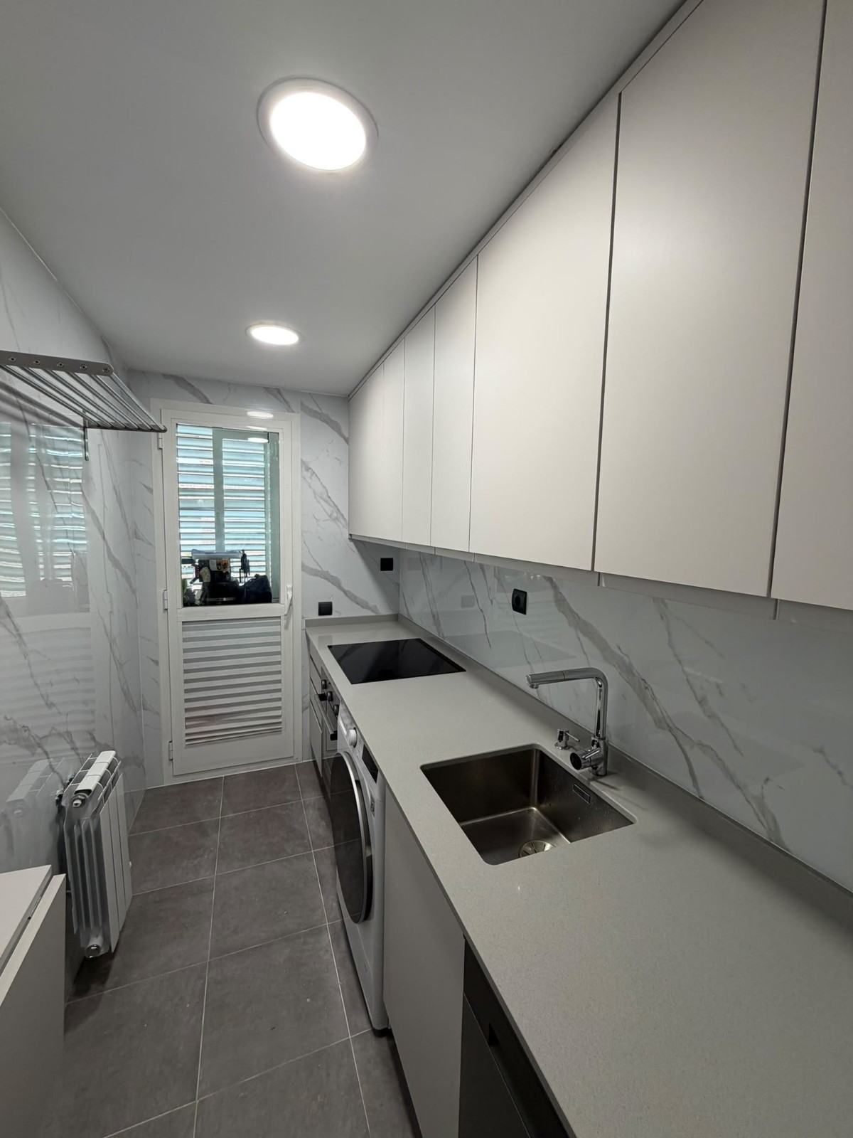 Cocina moderna, estrecha y blanca, con armarios empotrados, suelo de baldosas grises y luces de techo brillantes