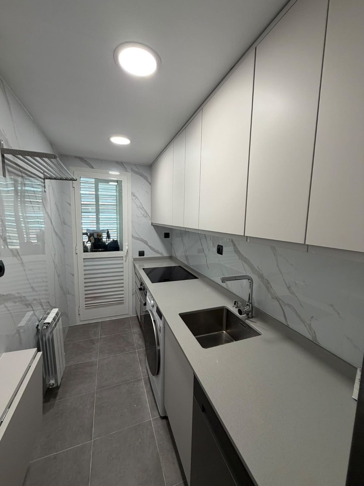 Cocina moderna y estrecha, con armarios blancos, suelo de baldosas grises y un fregadero junto a una ventana luminosa.