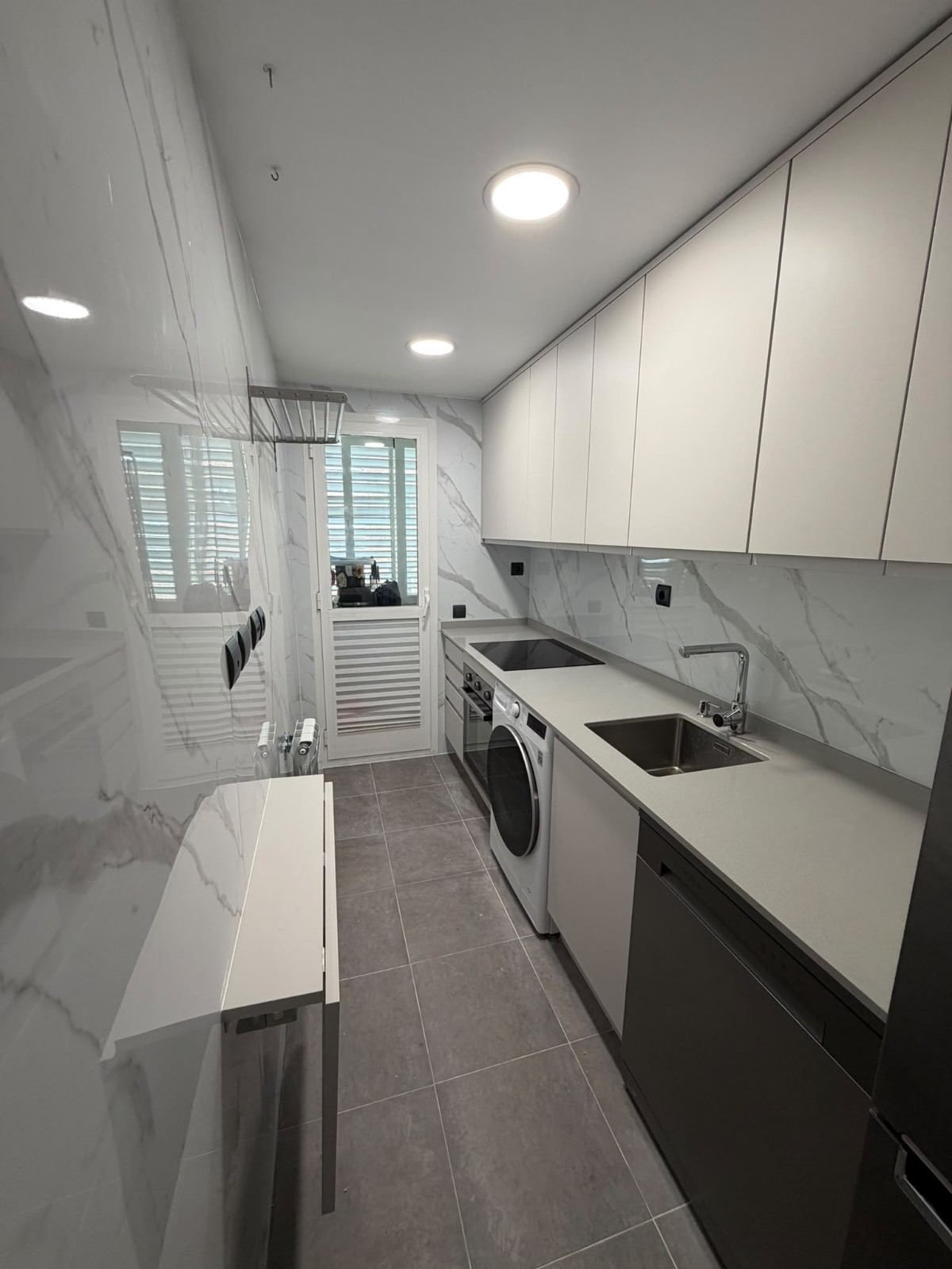 Cocina moderna y estrecha de estilo galera, con armarios blancos, salpicadero efecto mármol, suelo gris y una puerta con ventanas al fondo.