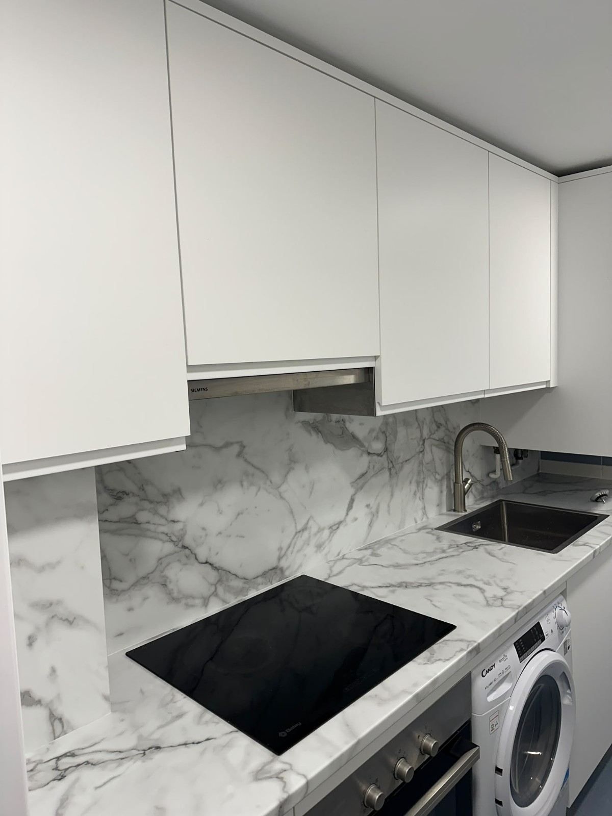 Cocina moderna con armarios blancos, salpicadero de mármol, encimera negra, fogones, fregadero y lavadora.
