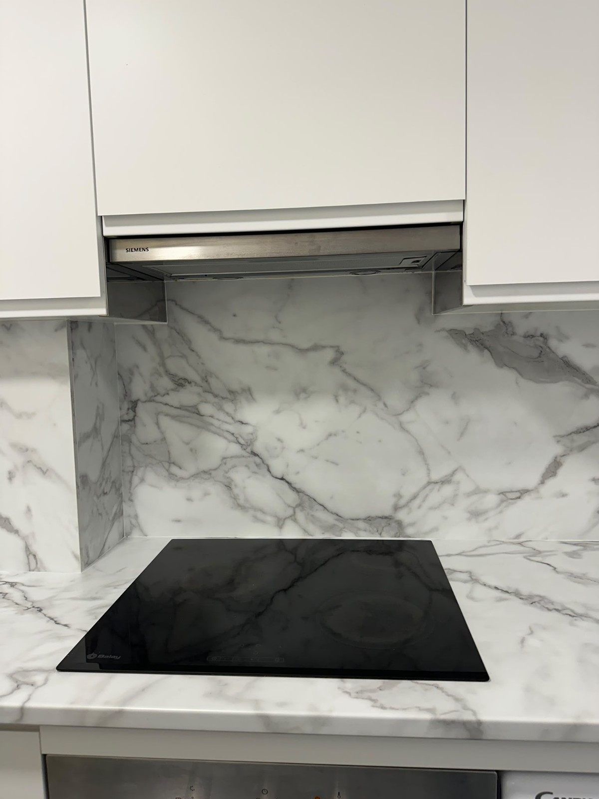 Cocina moderna con salpicadero de mármol, armarios blancos y una placa de inducción negra.