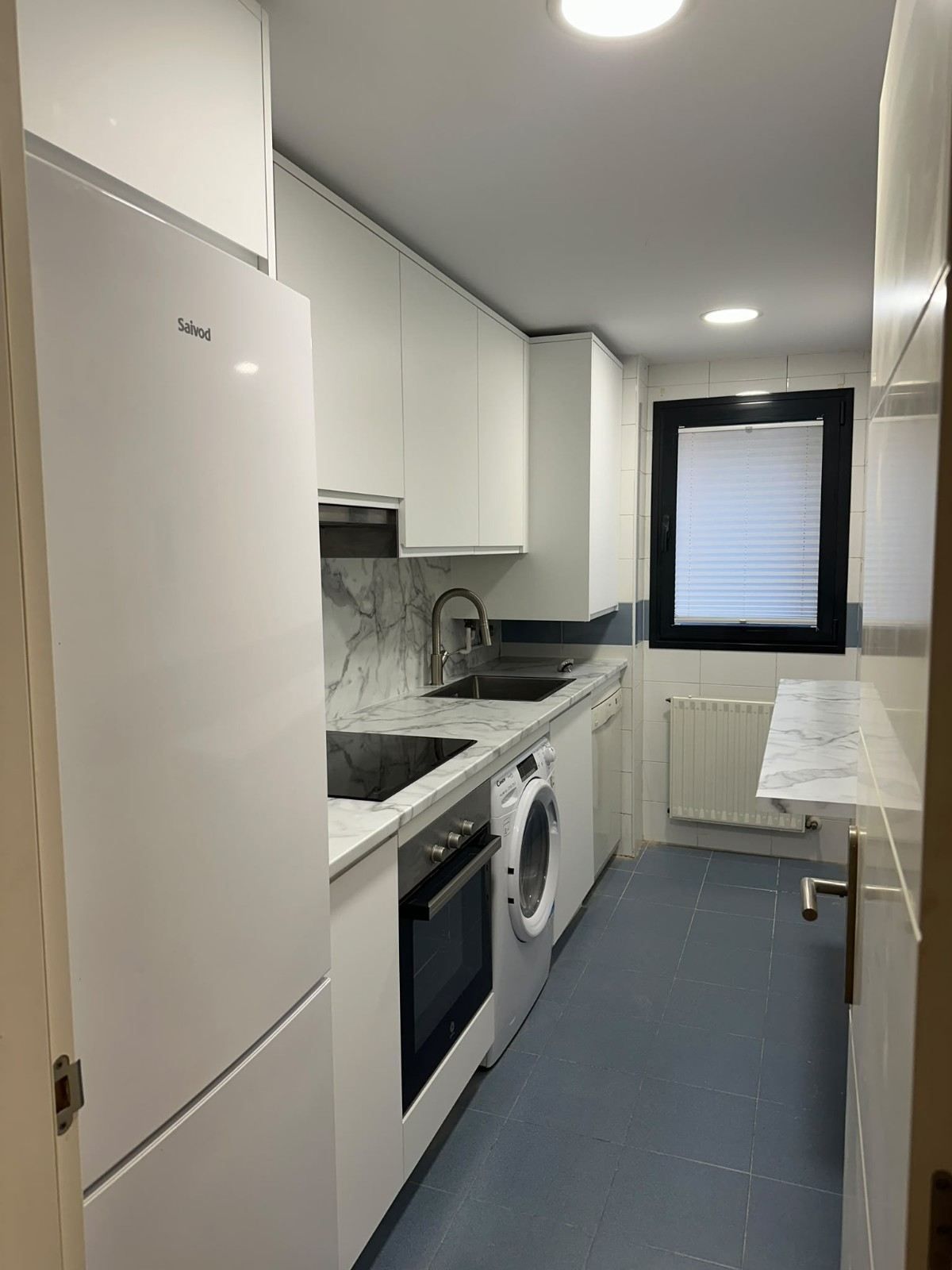 Cocina estrecha y blanca con suelo azul, lavadora, horno, fregadero y una ventana al fondo.