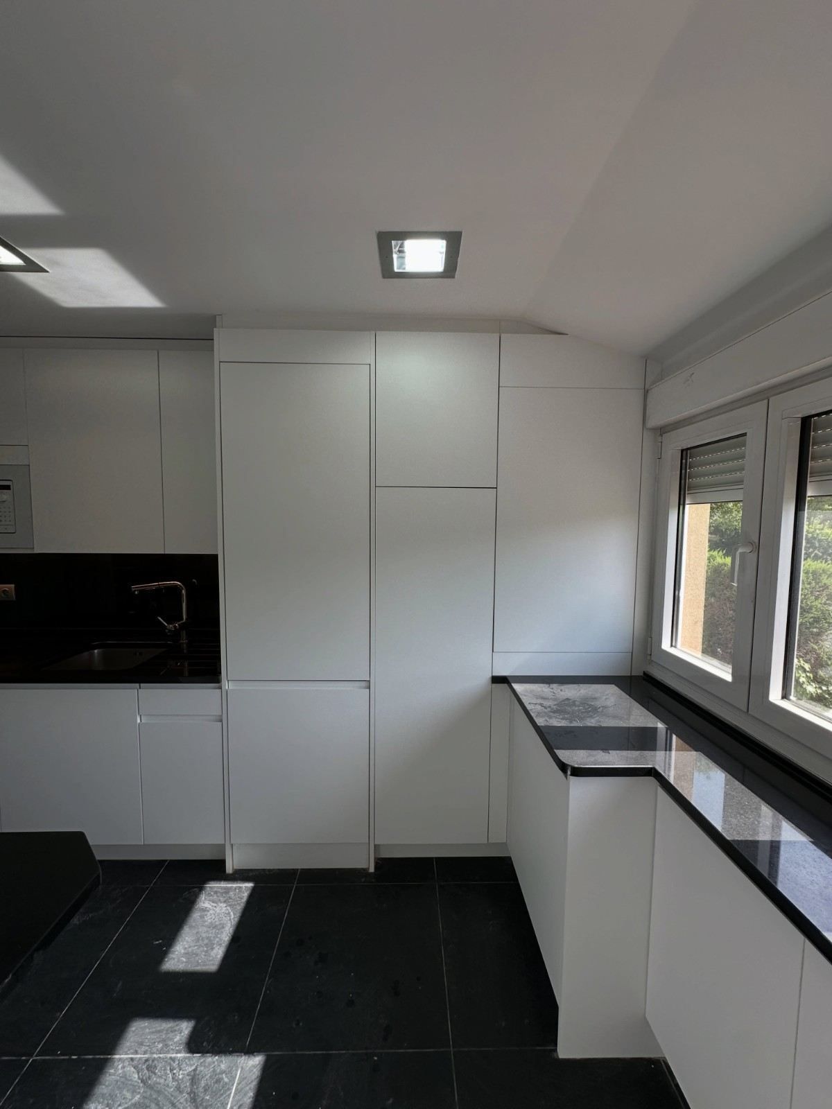 Cocina moderna blanca con encimeras negras, suelo de baldosas negras y grandes ventanas por las que entra la luz del día