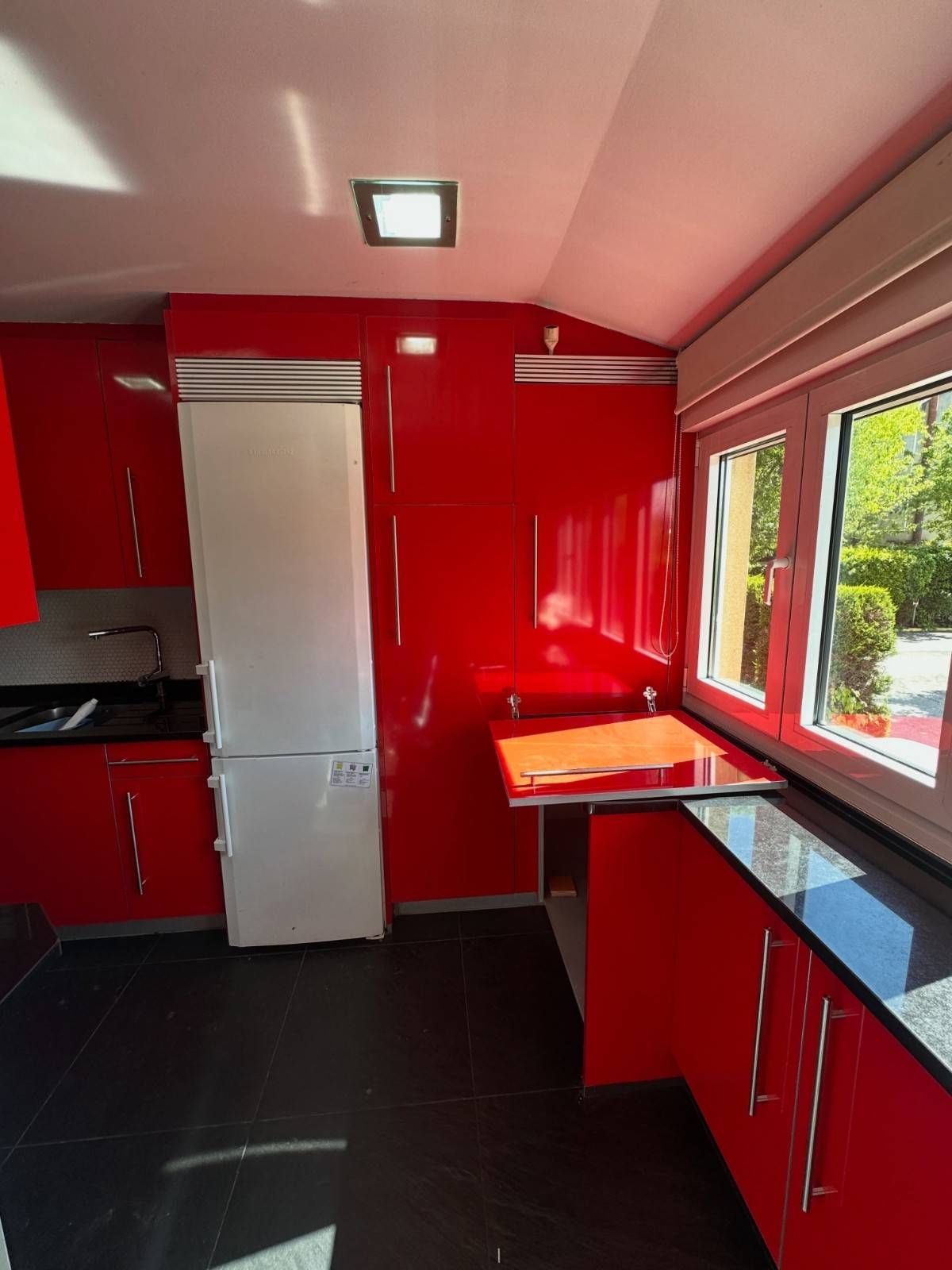 Cocina de color rojo intenso con refrigerador blanco, encimeras negras y una gran ventana bañada por el sol