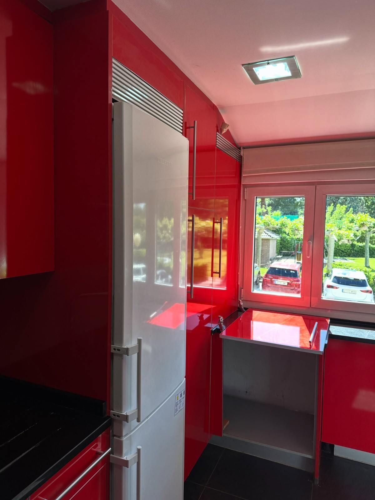 Cocina de color rojo intenso con un frigorífico blanco, una encimera negra y una ventana con vistas al exterior.