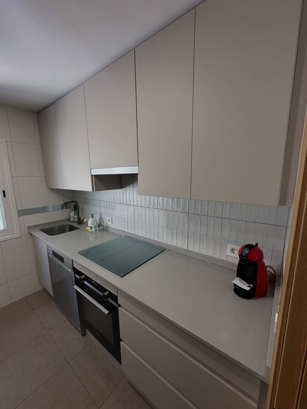 Cocina moderna con armarios blancos, encimera gris, placa de cocina, horno, fregadero y cafetera roja.