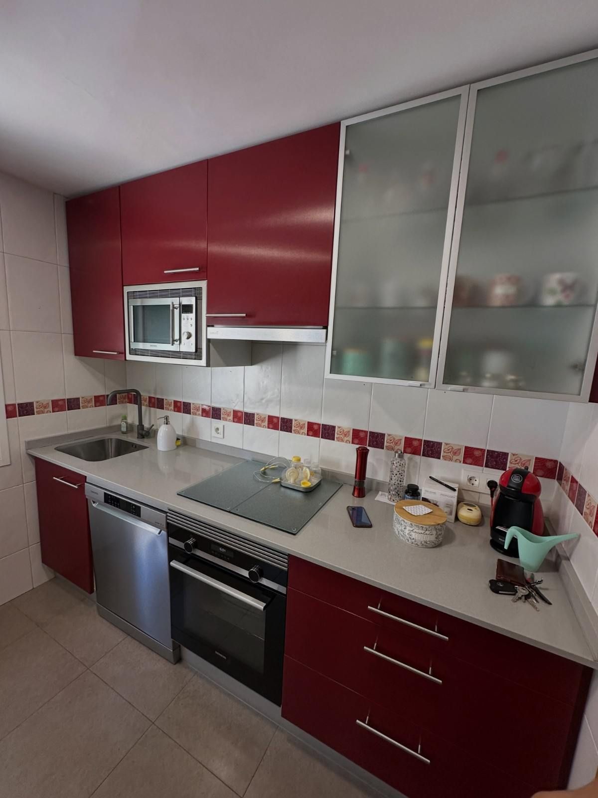 Cocina moderna con armarios rojos, encimeras blancas, horno empotrado, microondas y armarios superiores con puertas de cristal