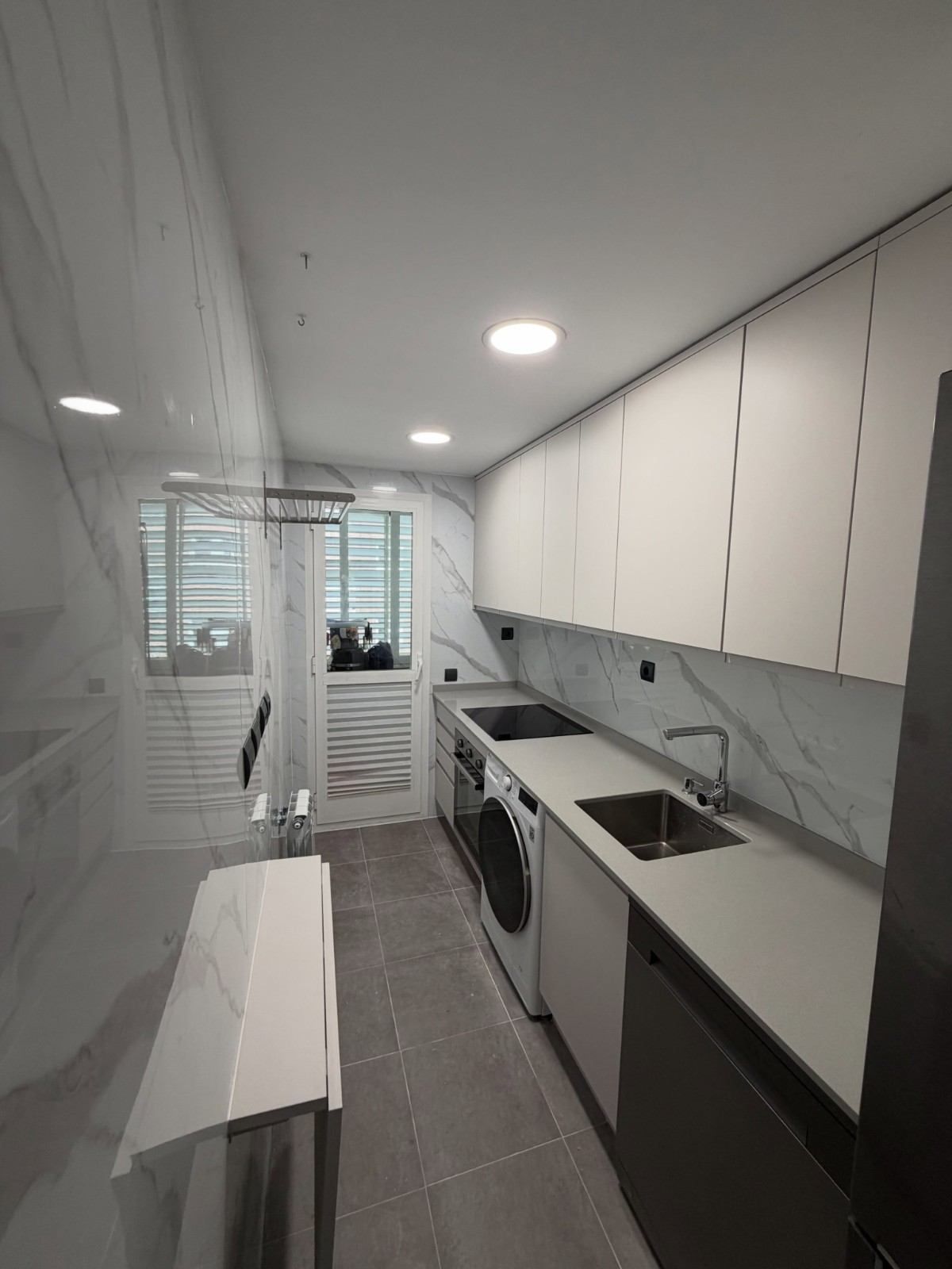 Cocina moderna y estrecha con armarios blancos, suelo de baldosas grises y una lavadora y secadora apiladas junto a una ventana.