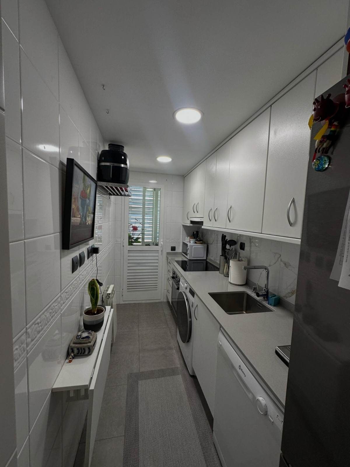 Cocina estrecha y blanca tipo pasillo, con armarios, fregadero, cocina y una alfombra gris alargada.