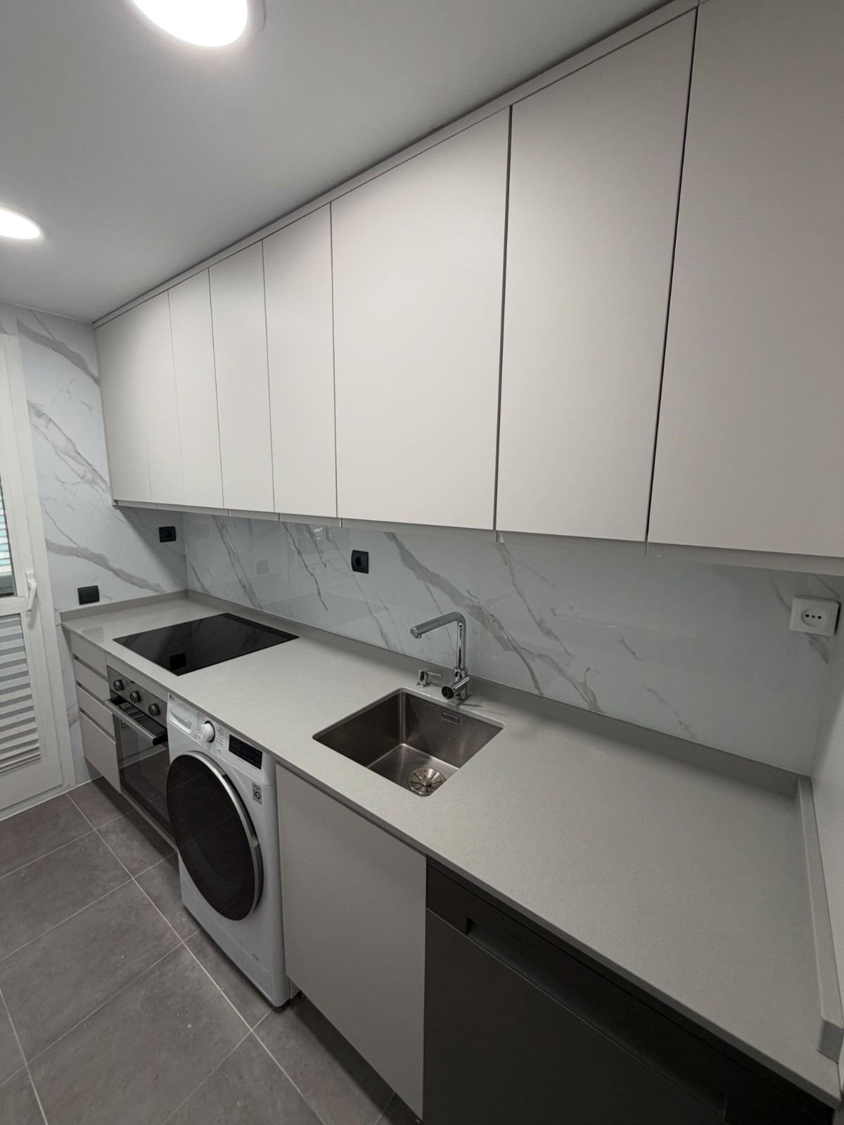 Cocina blanca moderna de estilo galera, con armarios superiores, encimeras efecto mármol, fregadero, lavavajillas y lavadora