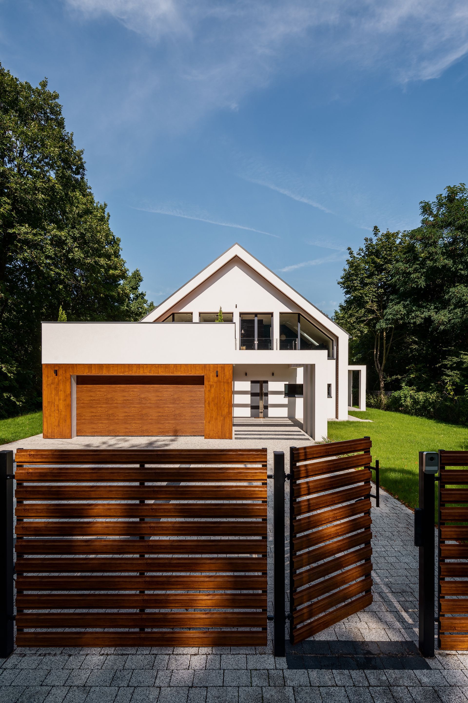 Maison blanche moderne avec des touches de bois, derrière une clôture et un portail en bois foncé, sous un ciel bleu.