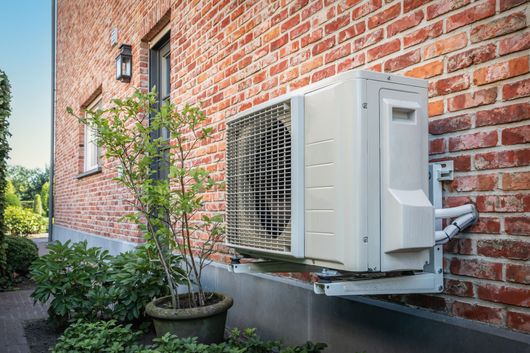 Unité de climatisation extérieure sur une maison en briques rouges avec des arbustes et un petit arbre.