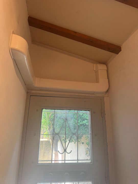 Climatiseur mural beige au-dessus d'une petite fenêtre à barreaux dans un coin de la pièce.