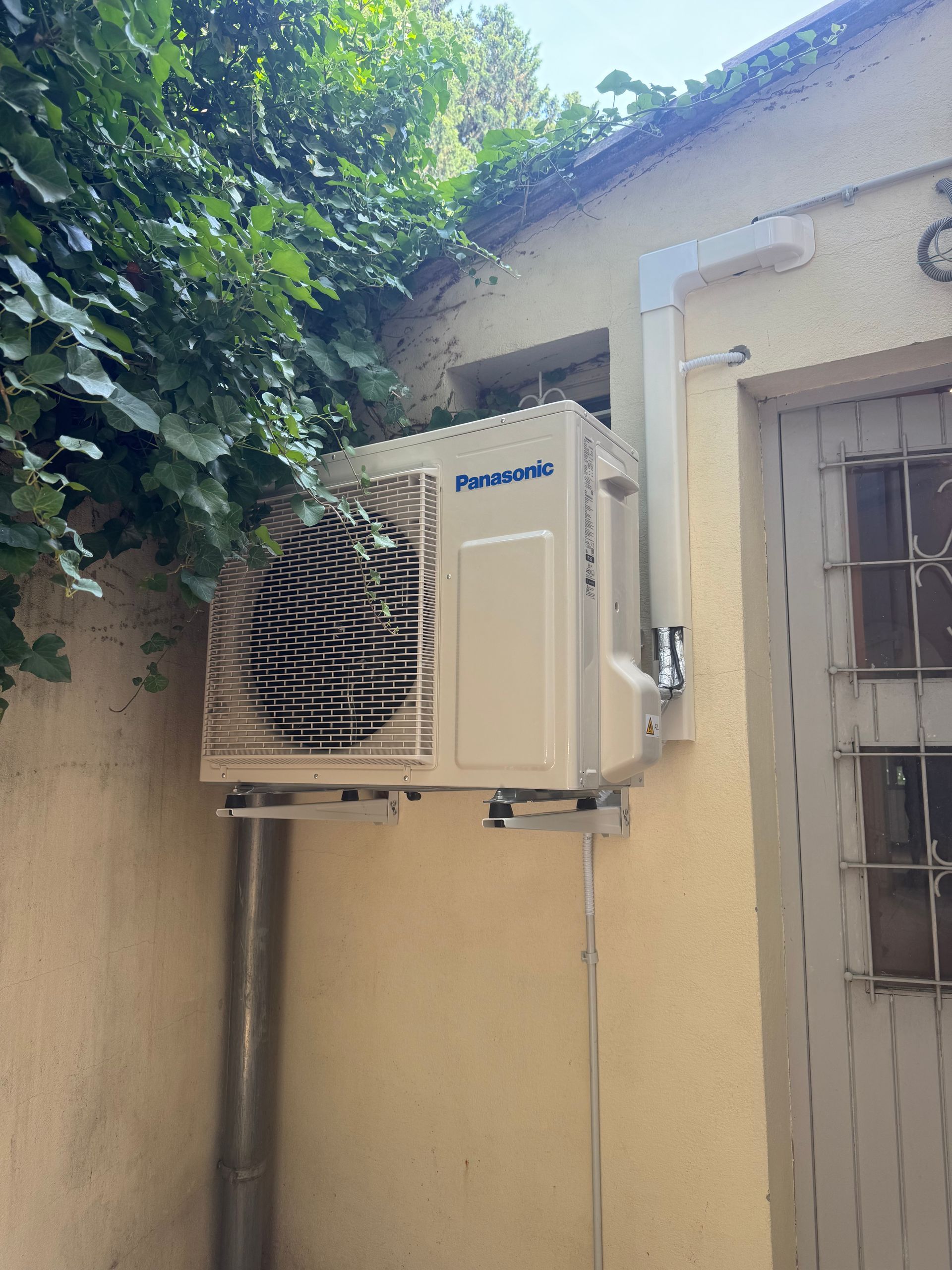 Unité de climatisation murale extérieure sur une maison beige, à côté d'une fenêtre à barreaux.