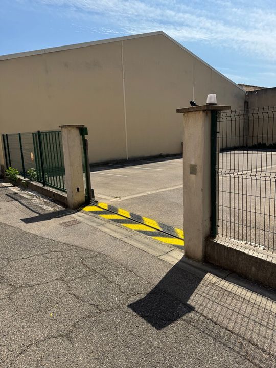 Portail ouvert donnant sur un bâtiment beige avec des ralentisseurs jaune et noir.