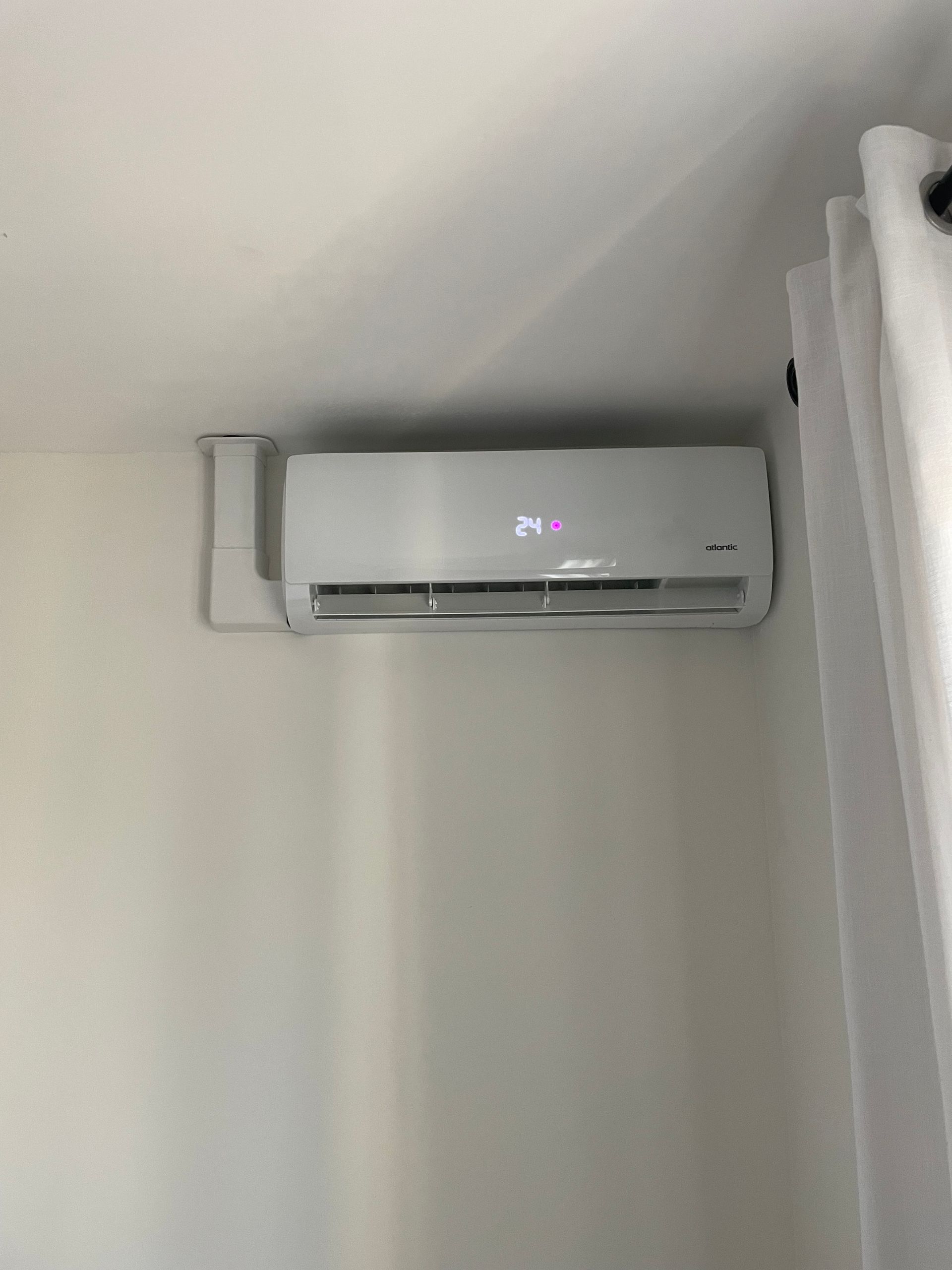 Climatiseur mural blanc au-dessus d'une fenêtre à rideaux, dans un coin de la pièce.