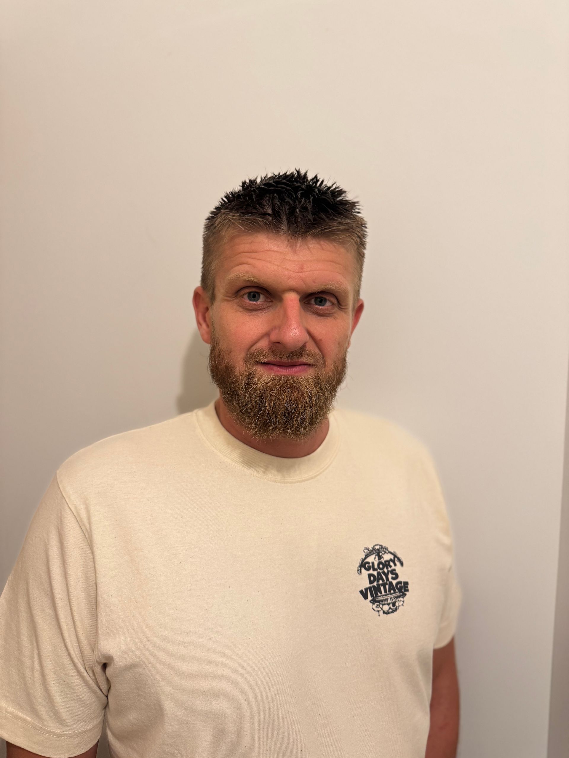 Un homme portant un t-shirt crème se tient debout contre un mur beige uni, esquissant un sourire.