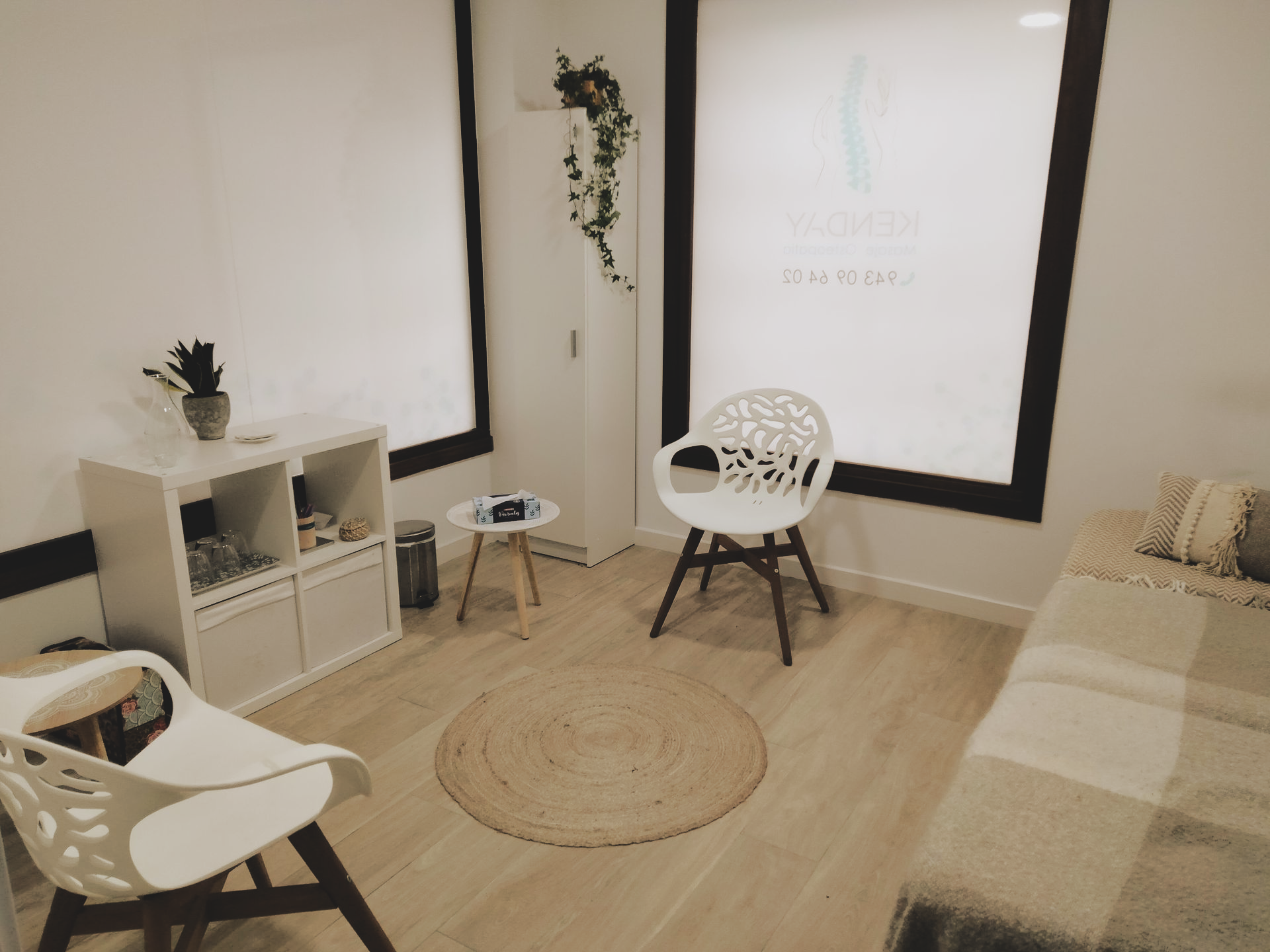 Un espacio de oficina lleno de luz con muebles blancos, una alfombra redonda y una mesa de masajes.