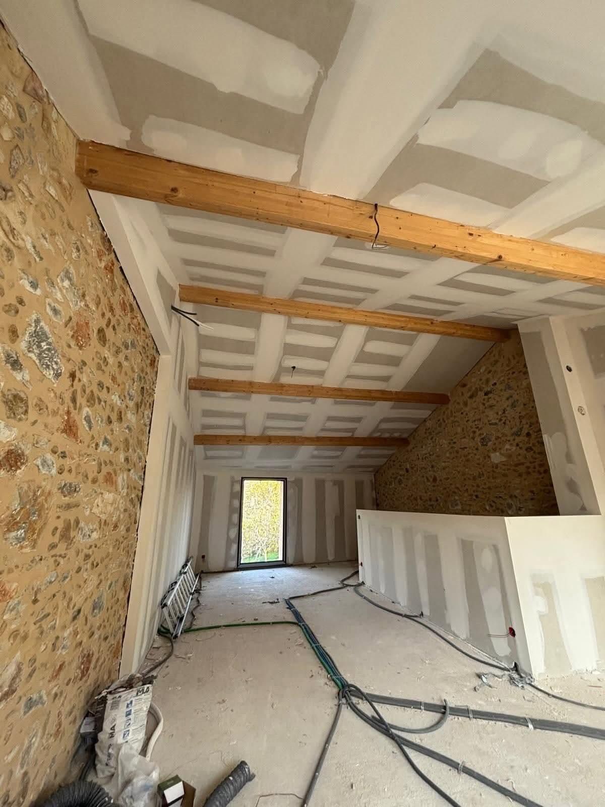 Pièce longue et étroite, inachevée, avec des murs en pierre, des poutres apparentes, une isolation blanche et des câbles au sol.