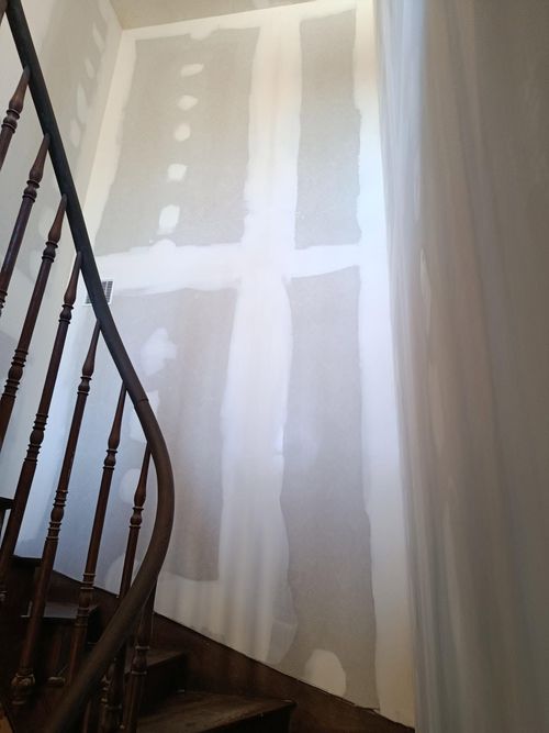 Mur de cage d'escalier avec des zones et des moulures recouvertes d'apprêt blanc, à côté d'un escalier en bois foncé.