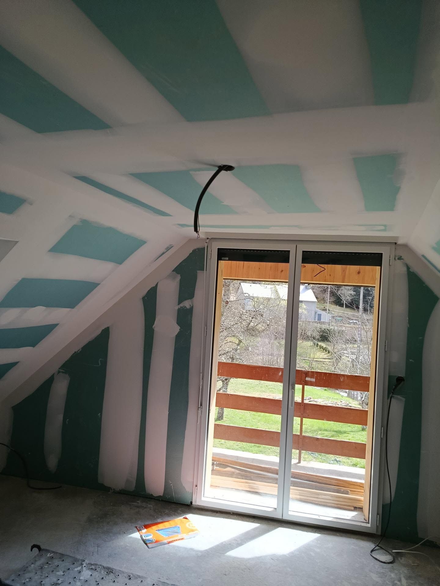 Pièce mansardée non aménagée avec des murs à rayures blanches et turquoise, un plafond mansardé et une porte coulissante en verre donnant sur l'extérieur.