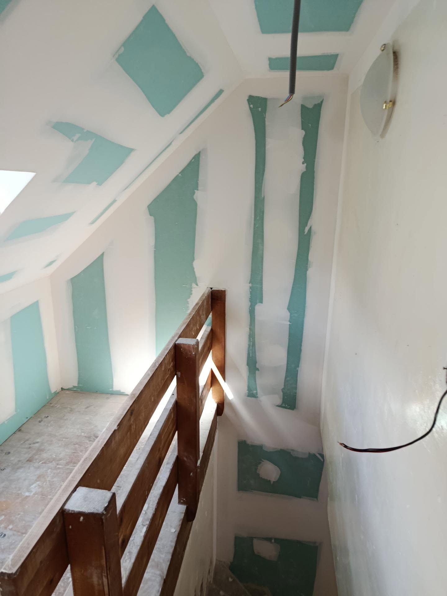 Couloir intérieur étroit en rénovation avec cloisons en Placo enduites, rampe en bois et luminaire apparent.