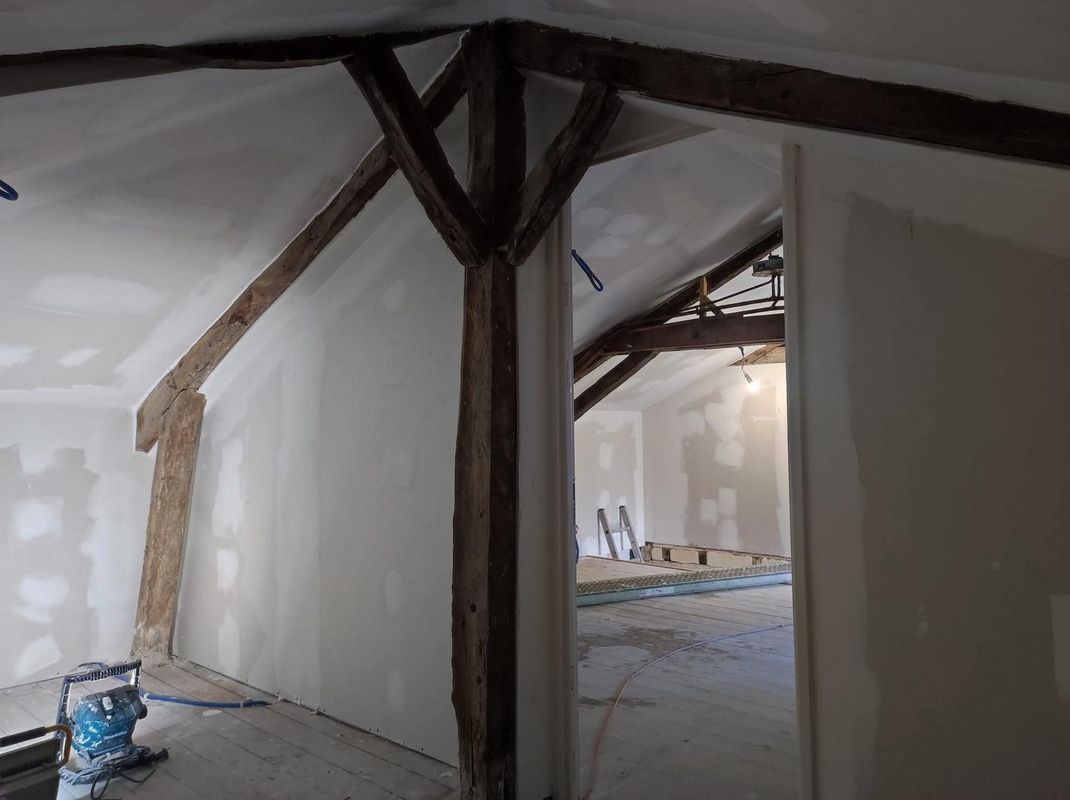 Intérieur d'une pièce partiellement rénovée avec poutres apparentes sombres, murs en plâtre blanc et une porte donnant sur une autre pièce.
