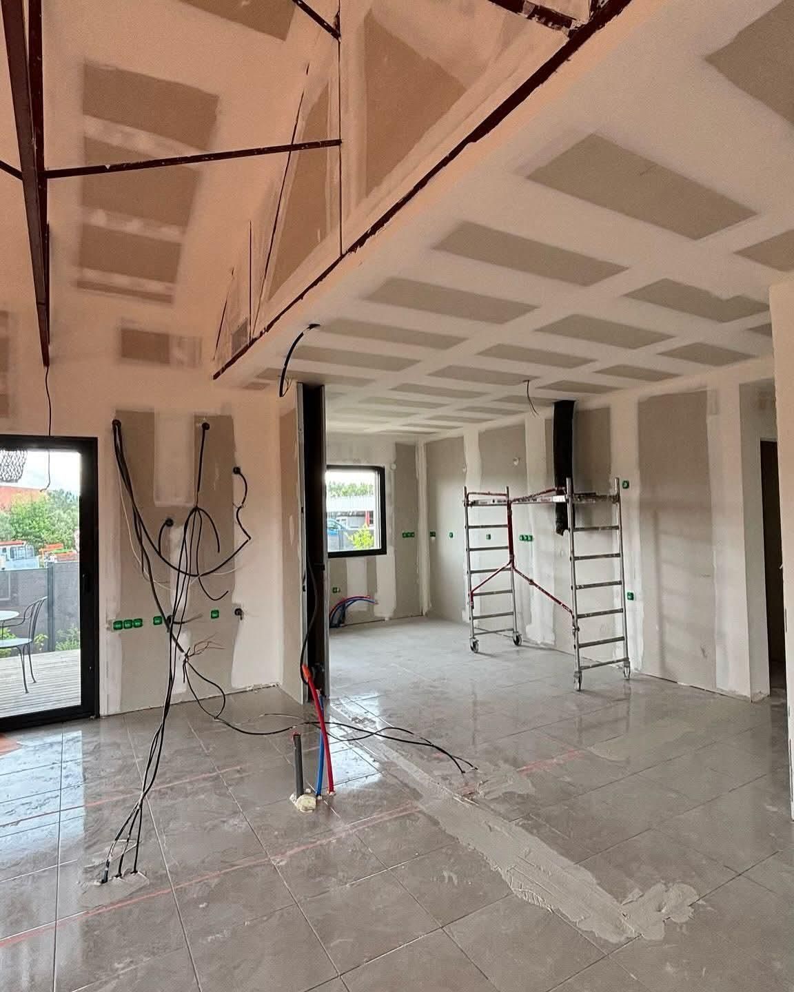 Intérieur d'une pièce en rénovation avec cloisons sèches, échafaudages, câbles apparents et une fenêtre laissant entrer la lumière du jour.