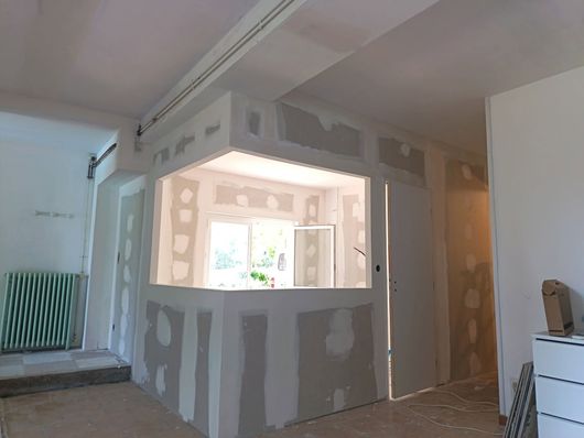 Intérieur partiellement rénové avec une cloison en plaques de plâtre grises et une grande fenêtre ouverte traversant.