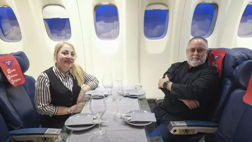 Dos personas sentadas en una mesa de avión, sonriendo, con bebidas y platos en una cabina con ventanas.