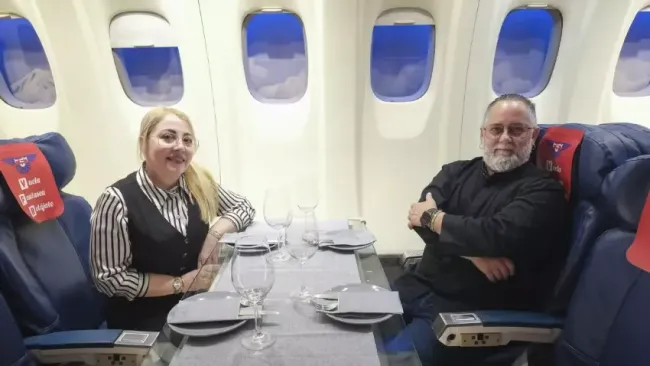 Dos personas sentadas en una mesa de avión, sonriendo, con bebidas y platos en una cabina con ventanas.