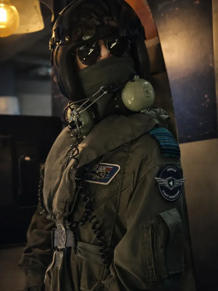 Piloto con traje de vuelo verde oliva, casco y gafas, de pie en el interior bajo una luz cálida.