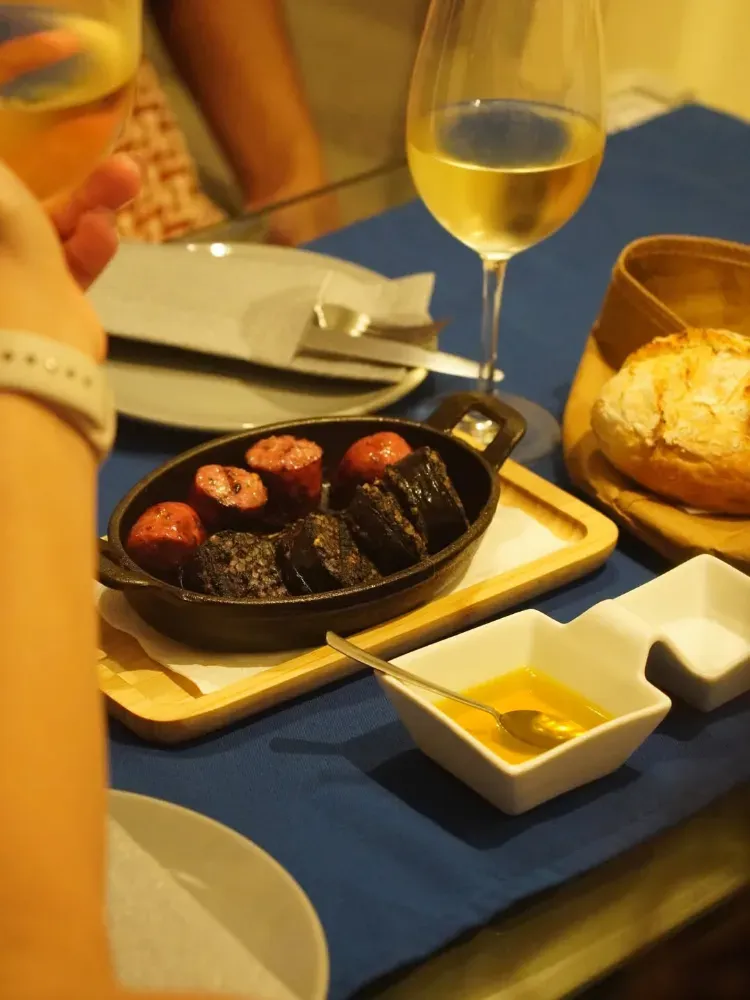 Vino y aperitivos sobre un mantel azul con un panecillo, salsas y un plato de albóndigas a la parrilla.