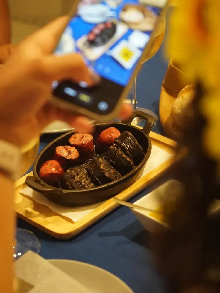 Una mano sosteniendo un teléfono inteligente sobre una mesa con una sartén negra llena de comida a la parrilla y fresas, con una iluminación interior cálida.
