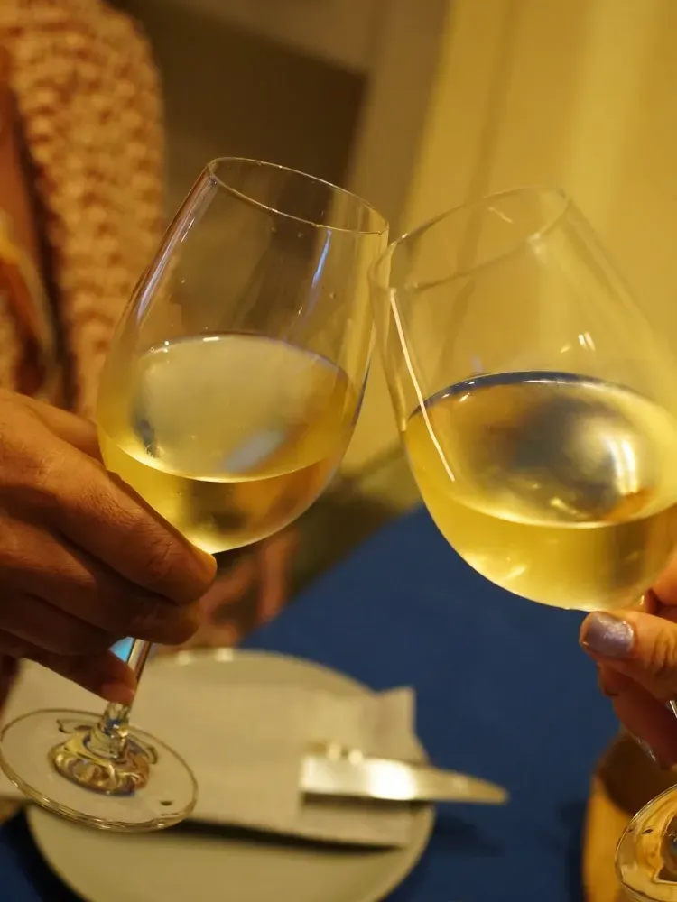 Dos manos alzando copas de vino blanco sobre una mesa durante un brindis.