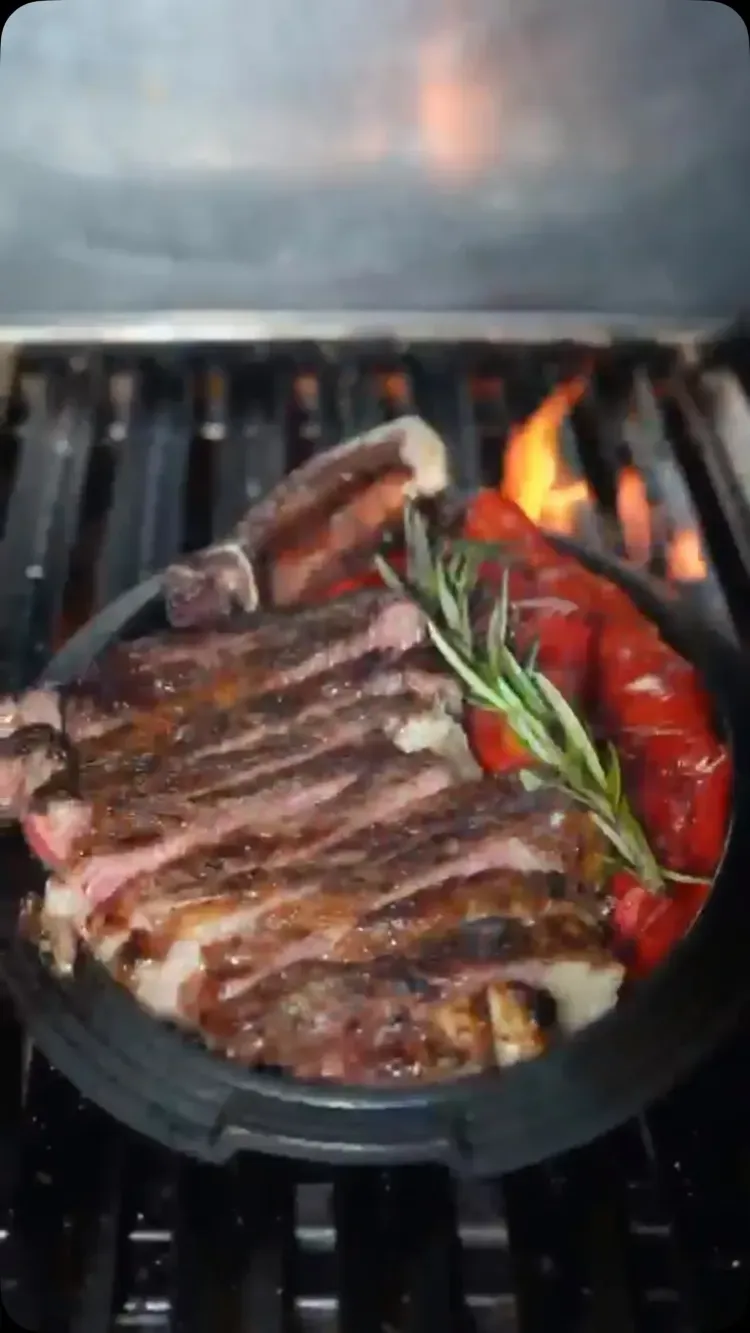 Filete a la parrilla con romero en una sartén de hierro fundido sobre la estufa, con tomates y llamas cerca.