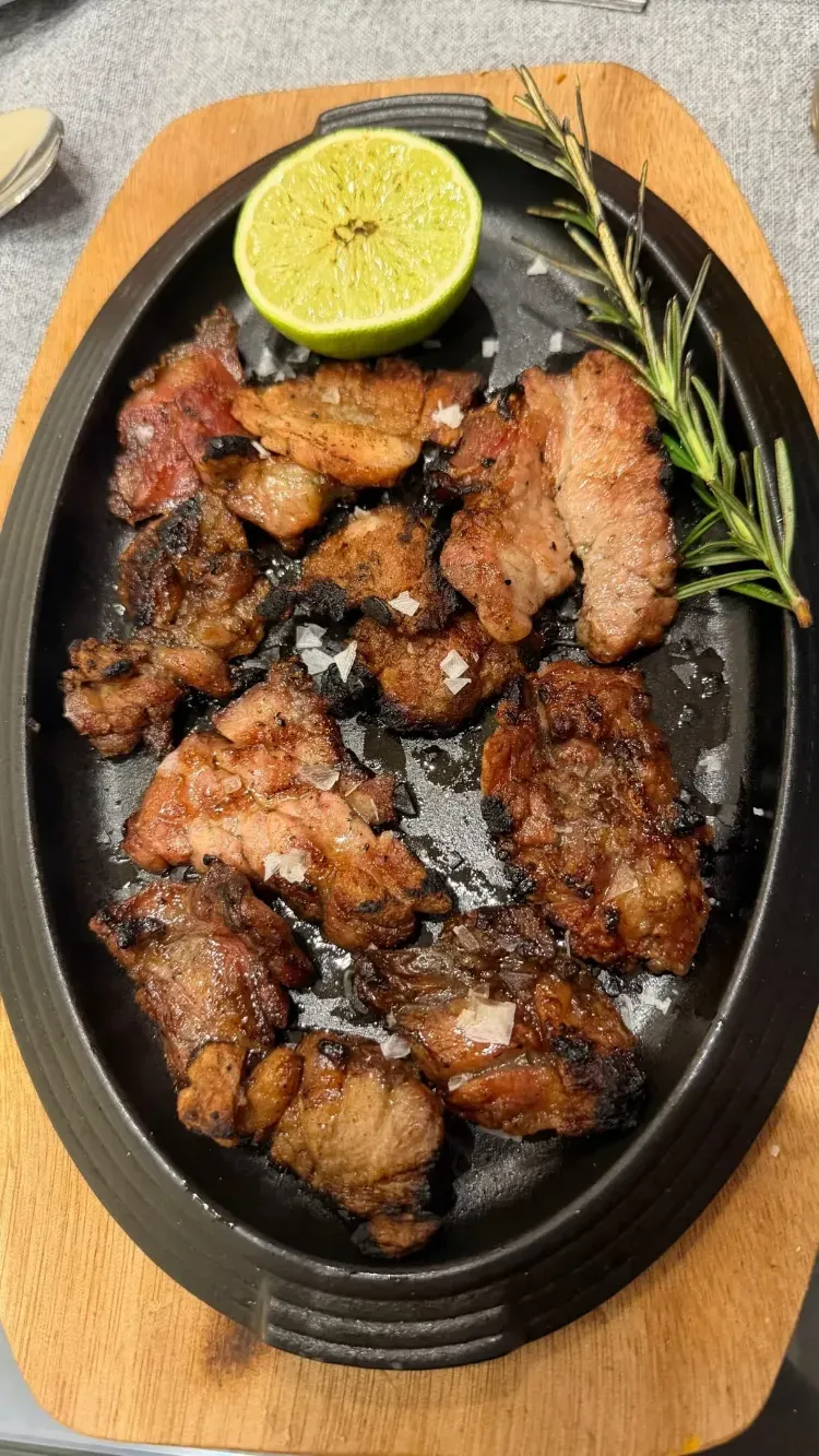 Carne a la parrilla en un plato negro con una rodaja de lima y romero sobre una tabla de madera.