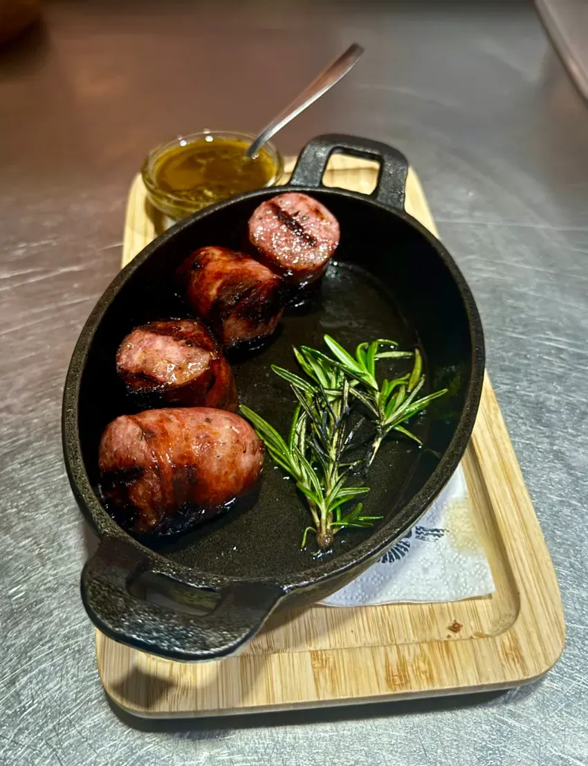 Sartén con carne sellada y romero en una bandeja de madera, con un pequeño frasco de salsa al lado.